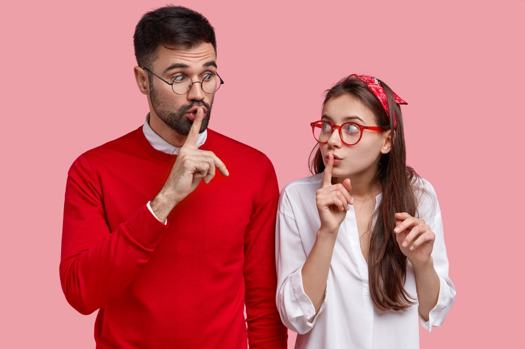 Dos personas haciendo un gesto de silencio, un hombre en suéter rojo y gafas, y una mujer con camisón blanco y gafas rojas, ambos con fondo rosa.