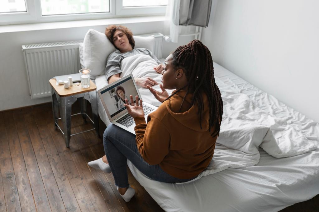 Una mujer sentada en una cama habla por videollamada con una profesional de la salud, mientras otra persona descansa en la cama cercana.