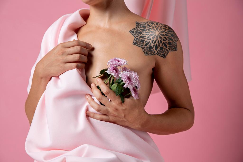 Persona sosteniendo flores, con un tatuaje en el hombro, vestida con una tela rosa frente a un fondo rosa.